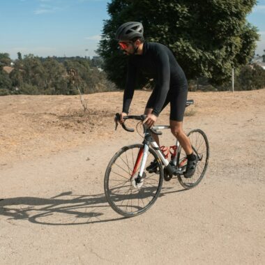 A cyclist wearing a helmet and sunglasses rides a bike on a sunny day in a natural setting.