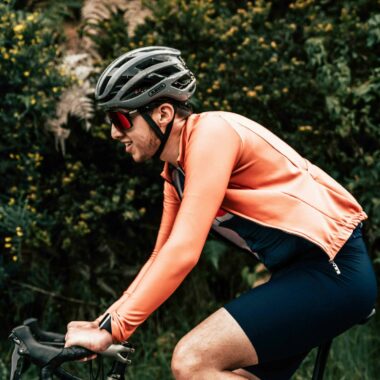 Cyclist in orange jacket and helmet biking through lush greenery, showcasing fitness and outdoor activity.